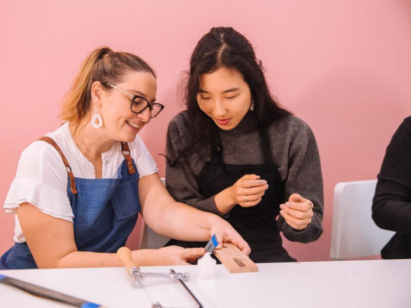Two women smiling as they help one another make jewellery